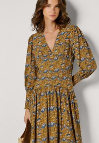 Sukienka w kwiatowy wzór w kolorze musztardowym z niebieskimi akcentami, z głębokim dekoltem w serek, długimi bufkowatymi rękawami i marszczonym dołem. Miękka faktura tkaniny.