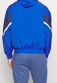 Man med en starkt blå huva-vindjacka med mörkblå och vita ärmaccent, i kombination med ljusblå byxor, sedd bakifrån.