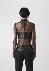 Top de encaje negro con mangas largas, diseño transparente, patrón floral y un ajuste estructurado con un panel negro en contraste en la cintura.