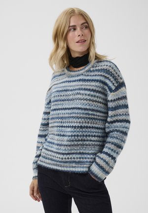 Femme aux cheveux blonds portant un pull duveteux rayé bleu, blanc et gris et un pantalon noir, regardant vers sa gauche sur un fond uni.