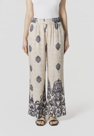 Mujer con pantalones anchos beige con patrones florales oscuros e intrincados y sandalias negras de punta abierta, posando frente a un fondo liso.