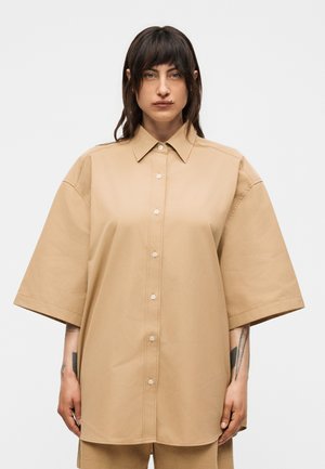 Γυναίκα με σκούρα μαλλιά μέχρι τους ώμους φοράει υπερμεγέθη μπεζ πουκάμισο με κοντά μανίκια και στέκεται μπροστά σε απλό λευκό φόντο.