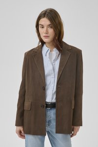 Jonge vrouw die een bruine suède blazer draagt over een blauw gestreept shirt en lichtgewassen spijkerbroek, staand tegen een effen grijze achtergrond.