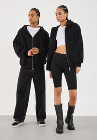 Hanorace negre cu fermoar, top alb scurt cu un design ajustat, pantaloni scurți negri până la genunchi și cizme negre până la genunchi cu accente din cataramă.