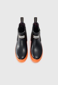 Bottes à chevilles en caoutchouc noir avec semelles orange, panneaux latéraux élastiques et une languette de tirage avec logo. Texture lisse avec un design à bout rond.