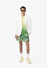 Hvid blazer, gul-grøn gradient polo, bladgrønne shorts, parret med gule sneakers, hvide sokker og sorte solbriller.