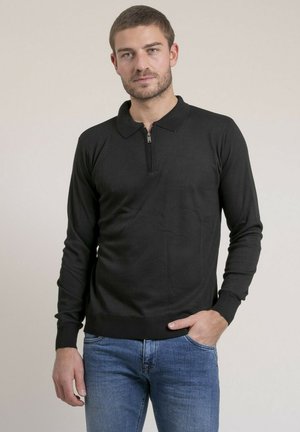 Homme aux cheveux courts et barbe, portant un polo noir à manches longues avec fermeture éclair et un jean bleu, debout avec une main dans la poche.