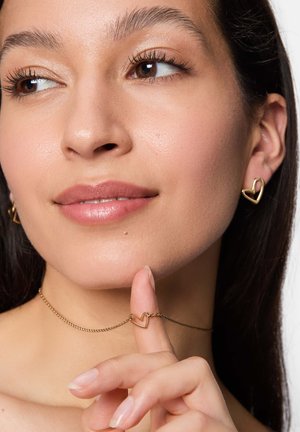 Goldene, herzförmige Ohrringe und eine Halskette an einem Model. Die Halskette hat einen kleinen Herzanhänger mit einer glatten, polierten Oberfläche.