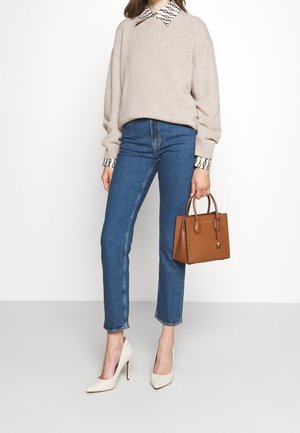Femme portant un pull beige par-dessus une chemise à motifs, un jean bleu, des talons blancs, tenant un sac à main en cuir marron sur fond blanc.