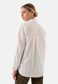 White button-up shirt with vertical pinstripes, long sleeves, and a subtle textured finish, worn over olive pants.