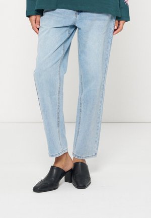 Personne portant un jean droit bleu clair et des mules en cuir noir à talons, debout devant un fond uni.