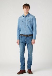 Lichtblauw denim shirt met twee borstzakken, gecombineerd met medium blauwe jeans. Een bruine riem en bordeauxrode schoenen maken de outfit compleet.