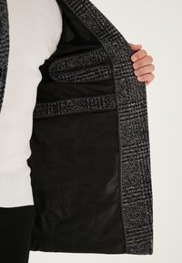 Abrigo texturizado en negro y gris forrado con tela suave; presenta bolsillos interiores negros visibles y costuras en contraste a lo largo de los bordes.