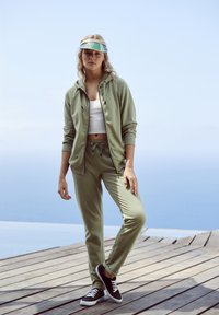 Olijfgroene zip-up jas met capuchon, bijpassende joggers en zwarte sneakers. Witte cropped top zichtbaar. Duidelijke vizier met regenboogband.