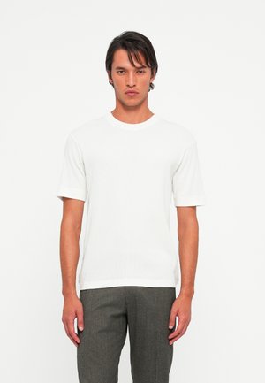 Young man with dark hair wearing a plain white short-sleeve t-shirt and gray pants, standing against a white background.