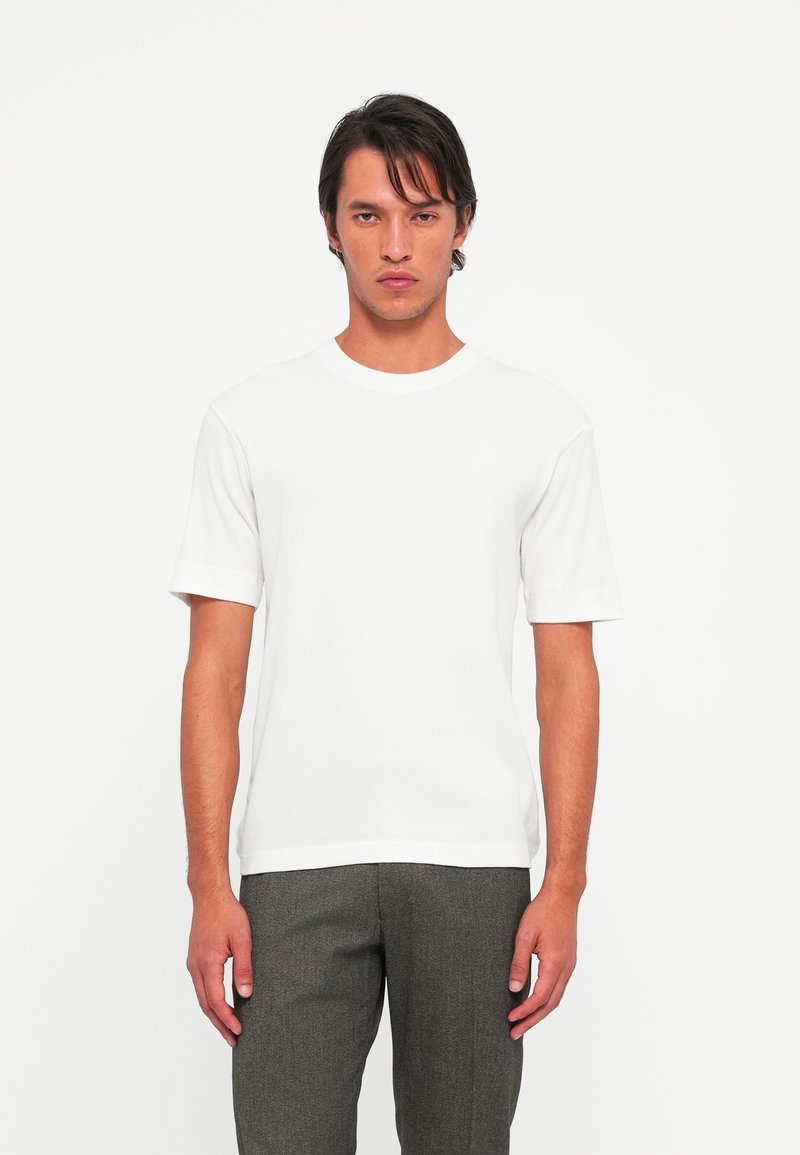 Young man with dark hair wearing a plain white short-sleeve t-shirt and gray pants, standing against a white background.