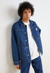 Jeansowa kurtka w ciemnym niebieskim kolorze z zapinaniem na guziki, dwiema kieszeniami na piersiach i małą łatą z logo. Połączona z białym t-shirtem i pasującymi dżinsami.