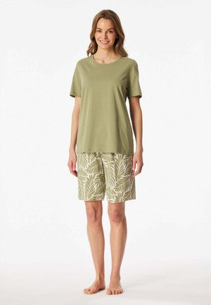 Donna in piedi a piedi nudi che indossa una maglietta verde oliva a maniche corte e pantaloncini beige con motivo di foglie verdi, sorridendo alla fotocamera.