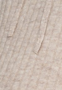 Beżowa dzianina z pionowym prążkowanym wzorem i cienkimi związaniami, nadająca miękki i przytulny wygląd.