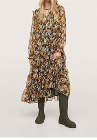 Nainen, joka on pukeutunut pitkään, läpinäkyvään kukkamekkoon, jossa on röyhelöitä, yhdistettynä oliivinvihreisiin polvipitkiin paksupohjaisiin saappaisiin, seisoo yksinkertaista taustaa vasten.