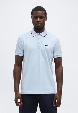 Uomo che indossa una polo BOSS azzurro chiaro con colletto a strisce nere e gialle, in piedi contro uno sfondo chiaro e uniforme.