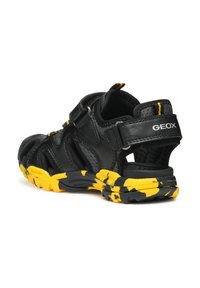 Sandalias negras y amarillas con parte superior de malla, correa ajustable y suela texturizada con un patrón de camuflaje. Logo de la marca en el talón.