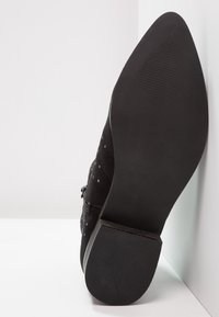 Zapato de ante negro con punta afilada, que presenta una suela texturizada y pequeños acentos decorativos. La suela es plana y suave, diseñada para proporcionar estabilidad.