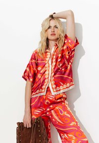 Ensemble coloré avec des motifs rouges et oranges, comprenant des designs floraux et ondulés ; chemise à manches courtes avec boutons et pantalon large assorti ; accompagné d'un sac marron.