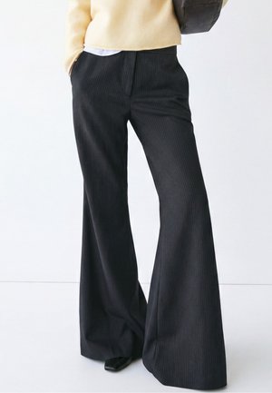 Persona che indossa pantaloni neri a righe ampie, maglione beige, con una mano in tasca e una borsa nera sulla spalla.