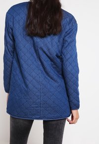 Blå vadderad jacka med diamantmönster, långa ärmar och rundad nederkant. Synliga sömmar och en texturerad yta.