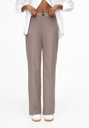 Person trägt hoch taillierte taupe Nadelstreifenhose mit Knopf und Reißverschluss, teilweise sichtbares weißes Hemd und weiße Sneakers mit Gummisohlen.