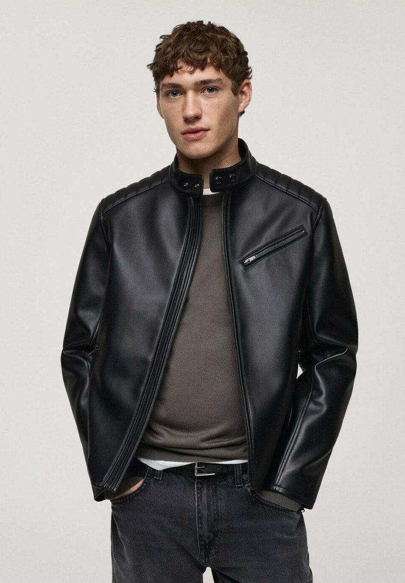 Giacca nera in finta pelle con colletto alla coreana, chiusura con zip e cuciture decorative. Abbinata a una camicia grigia e jeans scuri.