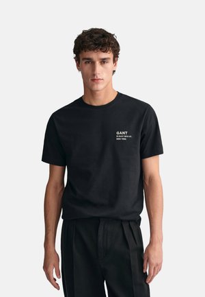 Camiseta de algodón negra con cuello redondo y mangas cortas. Presenta el texto del logo GANT en blanco y una dirección impresa en el área del pecho izquierdo.