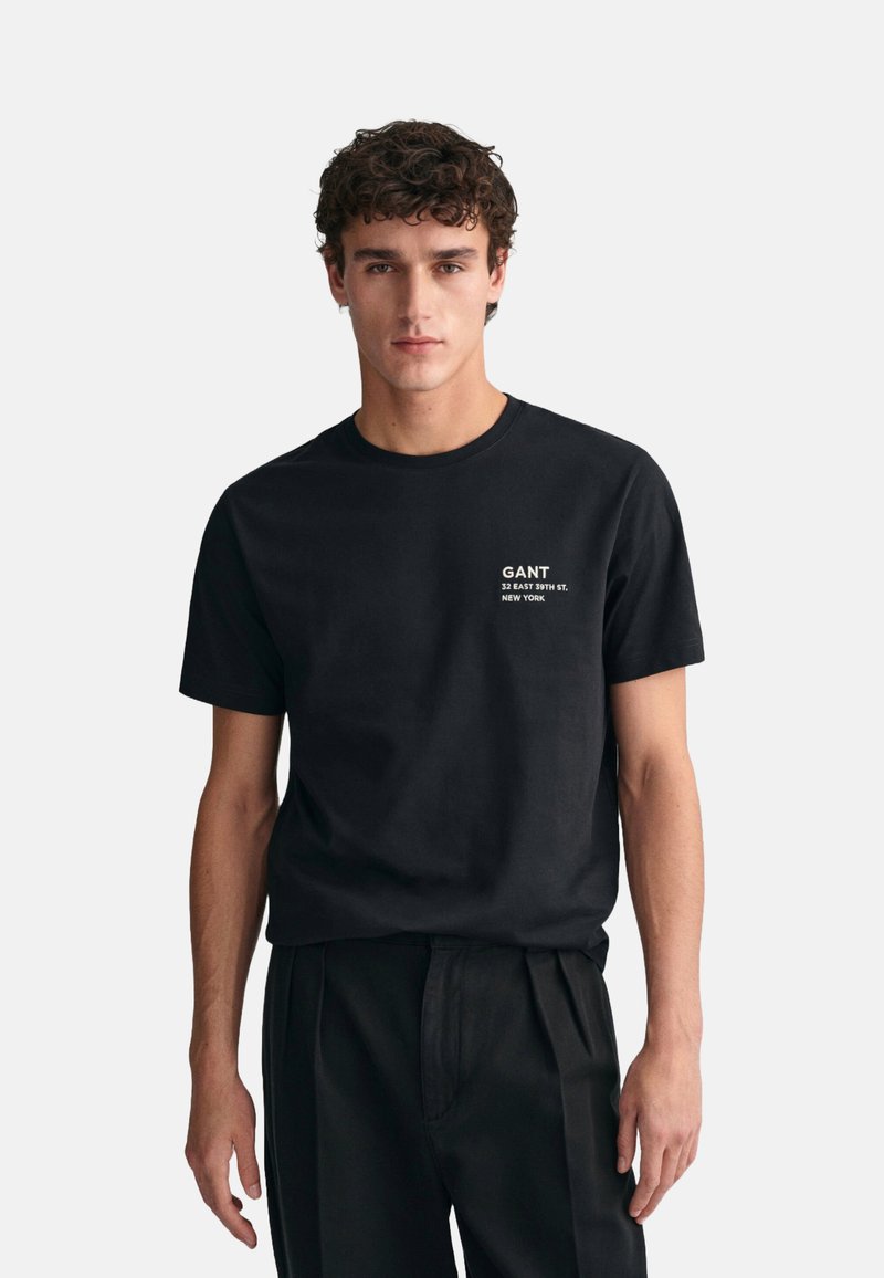Camiseta de algodón negra con cuello redondo y mangas cortas. Presenta el texto del logo GANT en blanco y una dirección impresa en el área del pecho izquierdo.