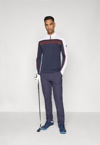 Marineblauwe langesleeve top met een ritssluiting kraag en bordeauxrode streep, gecombineerd met marineblauwe broek en blauwe schoenen, met een golfclub in de hand, met een witte handschoen aan.