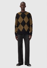 Männliches Model trägt einen braun-schwarzen Argyle-Pullover, schwarze gerade Jeans und schwarze Stiefel und steht vor einem schlichten Hintergrund.