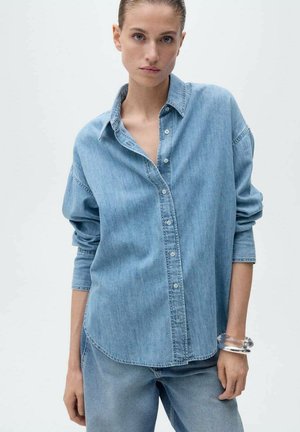 Helles blaues Denim-Hemd mit Knopfleiste, lockerer Passform und hochgekrempelten Ärmeln. Kombinieren Sie es mit passenden blauen Denim-Hosen. Transparenter Armreif am Handgelenk.