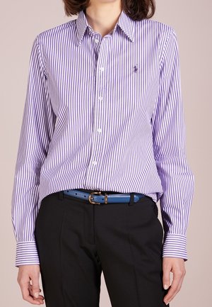 Osoba ubrana w koszulę na guziki w pionowe pasy w kolorze fioletowo-białym, wpuszczoną do czarnych spodni z niebieskim paskiem z złotą klamrą.