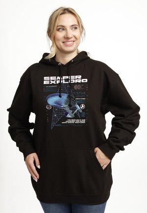 Kobieta w czarnej bluzie z kapturem z grafiką statku kosmicznego ze Star Treka i napisem „Semper Exploro, To boldly go where no one has gone before”, uśmiechająca się.