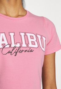 Růžové bavlněné tričko s odvážnými bílými blokovými písmeny, na nichž je napsáno "MALIBU" a "California" ve spojeném písmu pod tím. Měkká textura, kulatý výstřih.