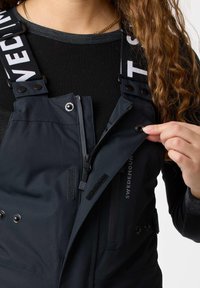 Persona che indossa una maglietta nera e una salopette blu navy con scritte bianche in grassetto, che allaccia un bottone a pressione sul davanti con zip e velcro visibili.