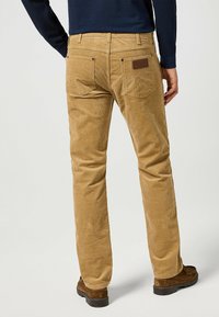 Beige ribfluwelen broek met een rechte snit, vijf zakken en een bruine leren patch op de achterste tailleband. Glad textuur en zichtbare stiksels.