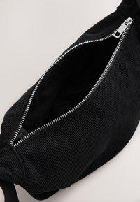 Zwarte corduroy tas met zilveren rits, voorzien van een geribbelde textuur en ruim interieur. Ontwerp met een enkele draagriem.