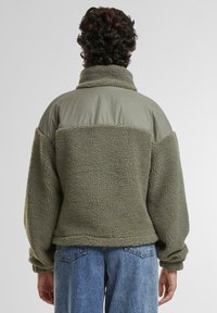 Olivgrön fleecejacka med hög krage, texturerad kropp och släta axelpartier. Avslappnad passform, sänkta axlar och uppkappade ärmar.
