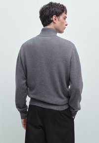 Uomo con capelli corti e scuri che indossa un maglione grigio a collo alto e pantaloni neri, girato leggermente di lato su uno sfondo semplice.