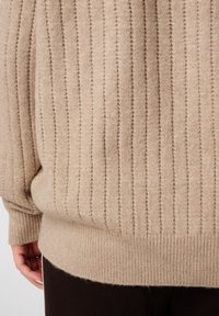 Pull en tricot beige à côtes avec une texture douce et un léger scintillement, une coupe décontractée et une large ceinture en bas.