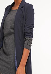 Blazer long bleu marine foncé à texture lisse, doté d'un col châle et de poignets retroussés sur un pull gris. Accents comprenant une doublure bleue contrastante.