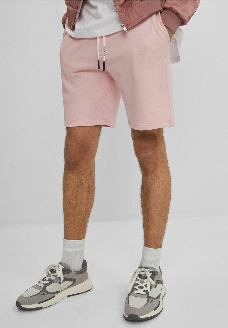 Homme portant un short à cordon de serrage rose clair, des baskets grises et blanches, des chaussettes blanches et une veste rose poussiéreuse partiellement visible.