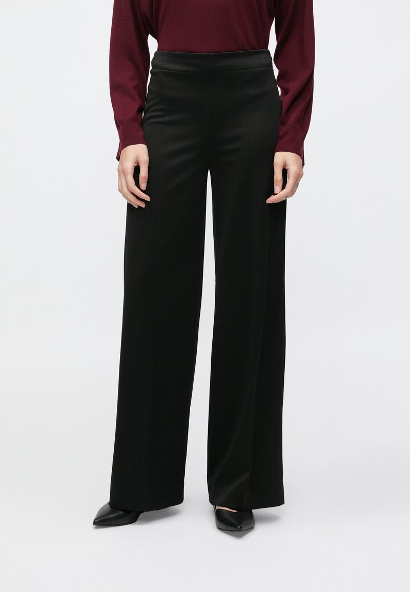 Pantaloni negri cu picioare largi, dintr-un material neted, cu talie înaltă și croială lejeră. Purtați cu un top grena cu mâneci lungi și pantofi cu toc negri.