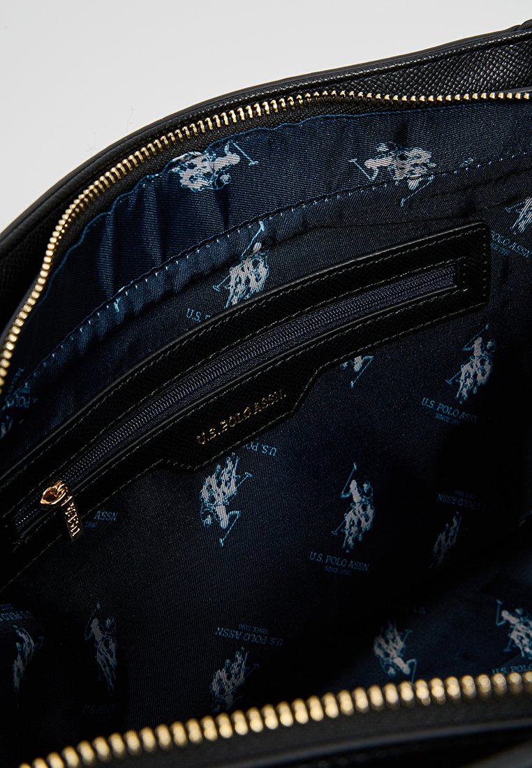 Interior de un bolso de mano de cuero negro con cremallera dorada, con forro azul oscuro que presenta un patrón repetido del logo de U.S. Polo Assn. en azul claro.